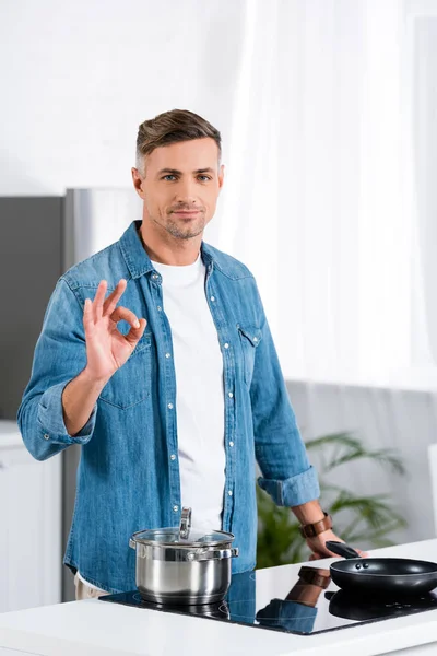 Guapo hombre adulto mostrando gesto bien y mirando a la cámara mientras está de pie en la cocina - foto de stock