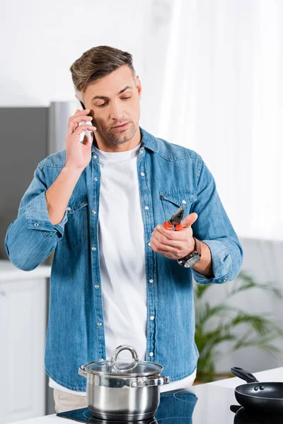 Hombre guapo hablando en el teléfono inteligente y sosteniendo alicates en la mano mientras está de pie en la cocina - foto de stock