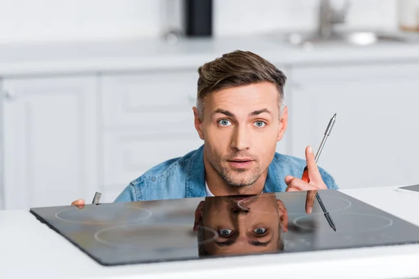 Hombre excitado reparando estufa eléctrica en la cocina, mostrando gesto de idea y mirando a la cámara - foto de stock