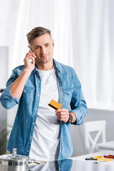 Усміхнений чоловік говорить на смартфоні і тримає кредитну картку на кухні — стокове фото