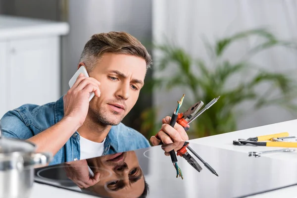 Man repairing wires of kitchen stove and talking on smartphone — Stock Photo