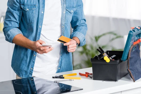 Vue recadrée de l'homme tenant smartphone et carte de crédit près de la boîte à outils — Photo de stock