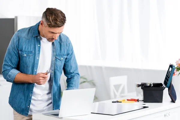 Adult man holding wrench and using laptop at kitchen — Stock Photo