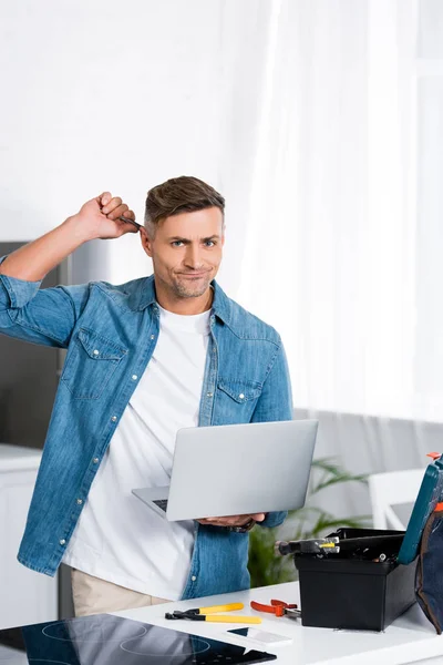 Hombre confundido sosteniendo portátil y rascando la cabeza con la herramienta - foto de stock