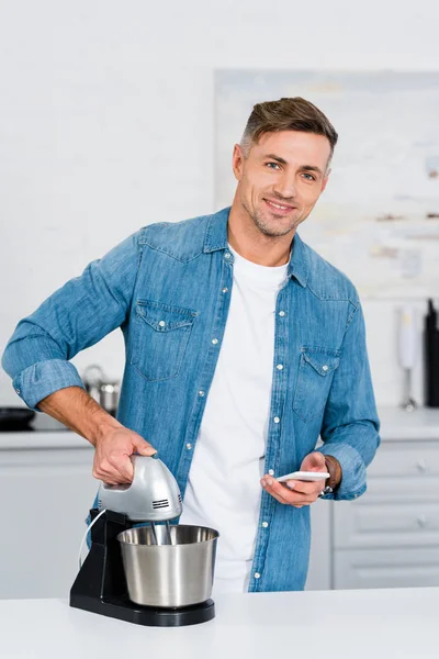 Homme adulte utilisant mélangeur de cuisine et smartphone tout en souriant à la caméra — Photo de stock