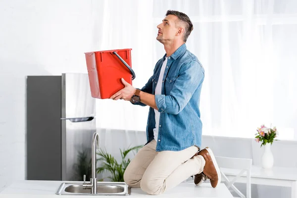 Uomo in piedi sulle ginocchia vicino lavello della cucina e tenendo secchio di plastica in mano mentre guardando soffitto — Foto stock