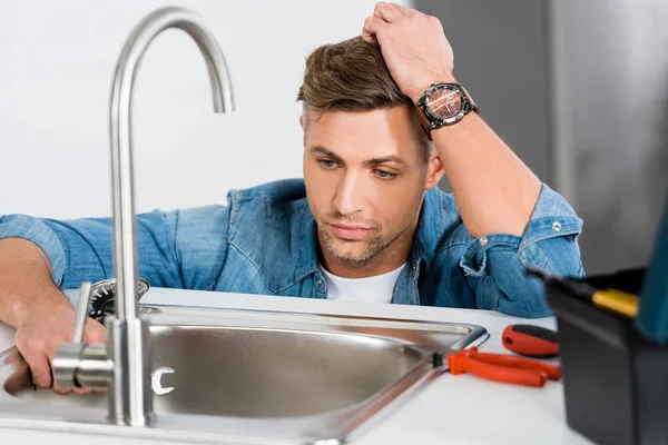 Sad handsome man looking at kitchen sink near tool box — Stock Photo