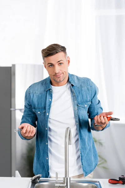 Homme confus avec des outils de réparation regardant la caméra tout en se tenant près de l'évier de cuisine — Photo de stock