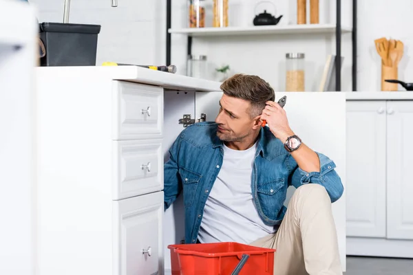 Verwirrter Mann sitzt auf dem Boden und repariert Küchenspüle — Stockfoto