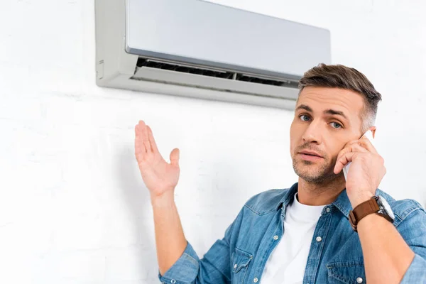 Hombre guapo hablando en el teléfono inteligente, mientras que de pie cerca de aire acondicionado - foto de stock