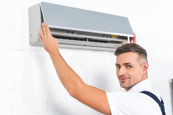 Beau réparateur vérifiant climatiseur et souriant à la caméra — Photo de stock