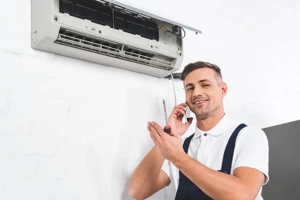 Hombre feliz hablando en el teléfono inteligente durante la reparación de aire acondicionado - foto de stock