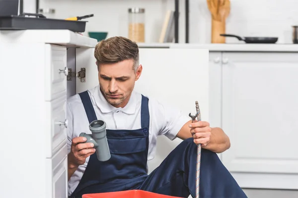 Thoughtful adult repairman sitting under sink and holding pipes for repairing at kitchen — Stock Photo