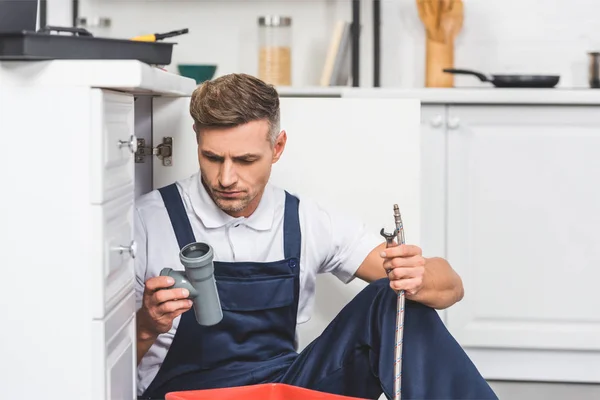 Reparador adulto reflexivo sentado debajo del fregadero y la celebración de tuberías para la reparación en la cocina - foto de stock