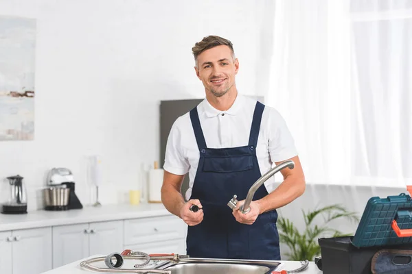 Lächelnder erwachsener Reparateur, der zum Reparieren den Küchenhahn abnimmt und in die Kamera schaut — Stockfoto