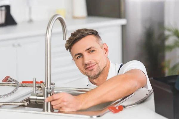 Riparatore adulto sorridente con chiave che ripara il rubinetto alla cucina e che guarda la macchina fotografica — Foto stock