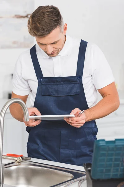 Вдумчивый взрослый ремонтник держит гаечный ключ и смотрит на цифровой планшет во время ремонта кухонного крана — стоковое фото