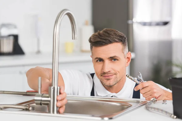 Smiling adult repairman holding pipe and spanner while repairing faucet at kitchen and looking at camera — Stock Photo