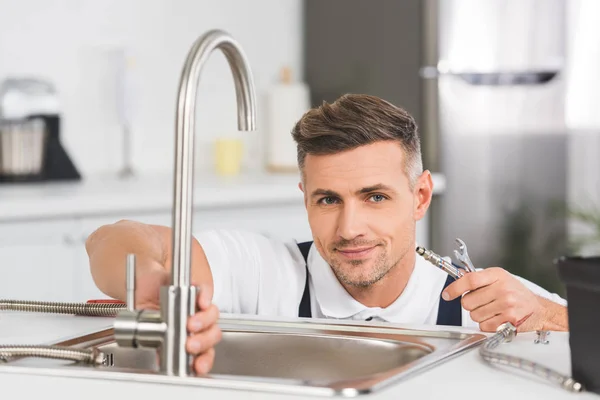 Riparatore adulto sorridente che tiene il tubo e chiave mentre ripara il rubinetto alla cucina e guarda la macchina fotografica — Foto stock