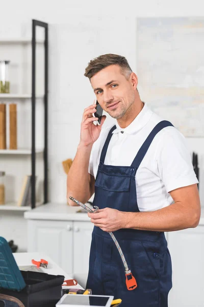 Улыбающийся взрослый ремонтник держит трубу для ремонта кухонного крана и смотрит в камеру, разговаривая на смартфоне — стоковое фото