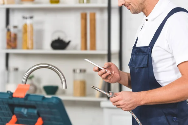 Ausgeschnittene Ansicht eines erwachsenen Reparateurs, der ein Rohr für die Reparatur von Küchenarmaturen und die Verwendung von Smartphones hält — Stock Photo
