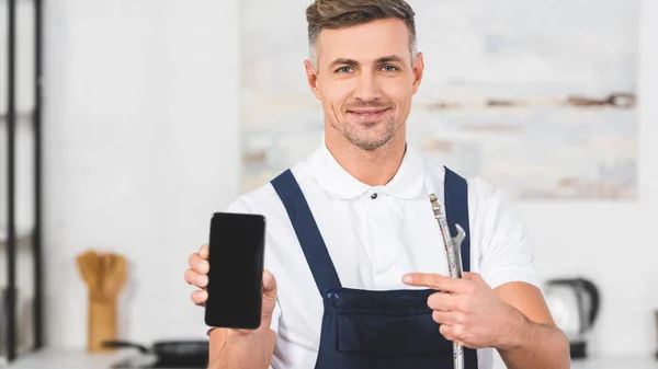 Smiling adult handyman holding pipe and pointing at smartphone with blank screen — Stock Photo