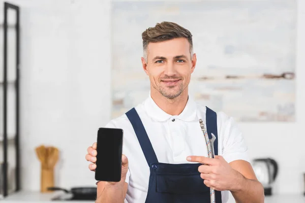 Sonriente hombre adulto sosteniendo la tubería y llave mientras que apunta a la pantalla en blanco del ingenio del teléfono inteligente - foto de stock