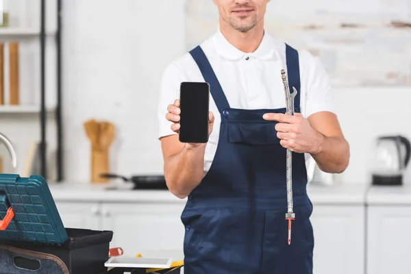 Abgeschnittene Ansicht eines lächelnden erwachsenen Mannes mit Pfeife und Schraubenschlüssel, während er auf den leeren Bildschirm des Smartphones zeigt — Stockfoto