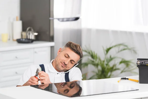 Adult repairman talking on smartphone while repairing electric stove at kitchen — Stock Photo