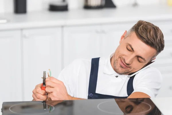 Reparador adulto hablando en el teléfono inteligente mientras que la reparación de la cocina eléctrica en la cocina - foto de stock