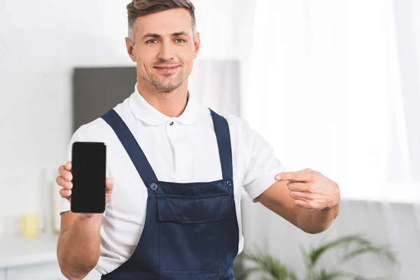 Sonriente reparador adulto sosteniendo el teléfono inteligente y apuntando con el dedo a la pantalla en blanco y mirando a la cámara - foto de stock