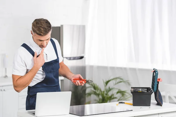 Thoughtful adult repairman looking at laptop and holding pliers while repairing electric stove — Stock Photo