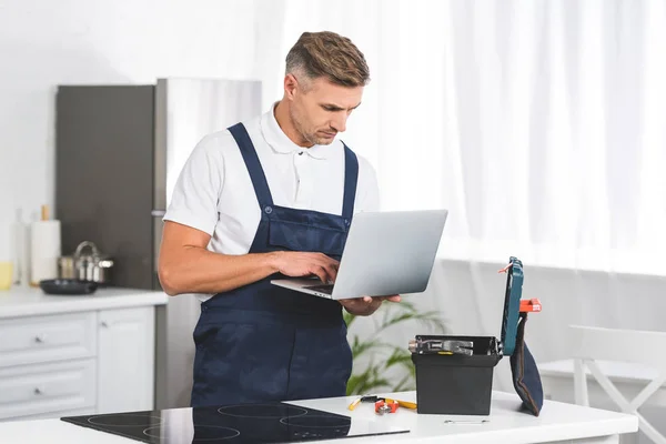 Reparador adulto pensativo usando el ordenador portátil mientras que repara la estufa eléctrica en la cocina - foto de stock