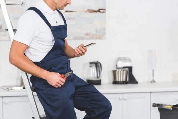 Обрізаний вид на ремонтника, який сидить на сходах тримаючи плоскогубці і використовуючи смартфон на кухні — стокове фото