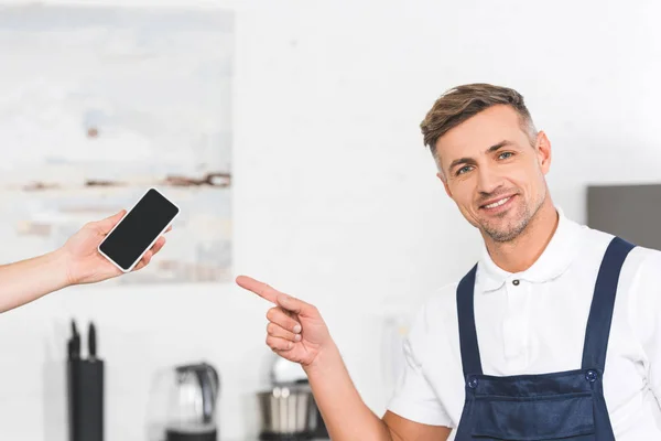 Vista parcial del teléfono inteligente con la mano y el reparador adulto sonriente apuntando a la pantalla en blanco - foto de stock