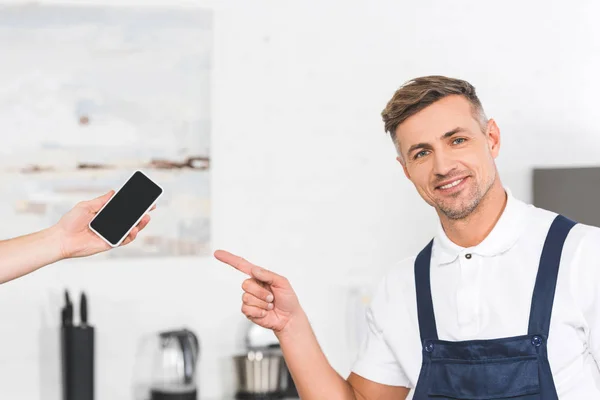 Teilansicht von Hand mit Smartphone und lächelndem erwachsenen Werkstattmitarbeiter, der auf leeren Bildschirm zeigt — Stockfoto