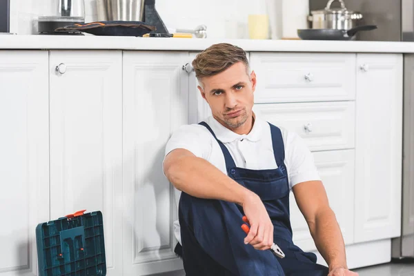 Upset adult repairman sitting and holding pliers at kitchen — Stock Photo