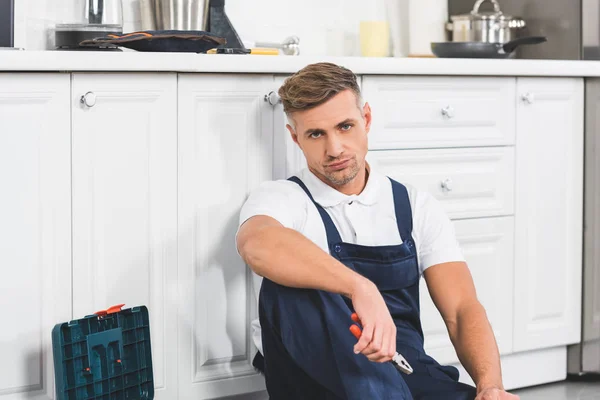 Perturbado reparador adulto sentado e segurando alicate na cozinha — Fotografia de Stock