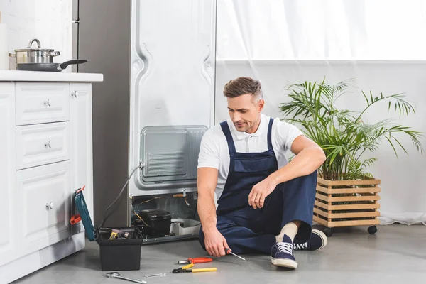 Уставший ремонтник, сидящий на полу и смотрящий на инструменты во время ремонта холодильника — стоковое фото