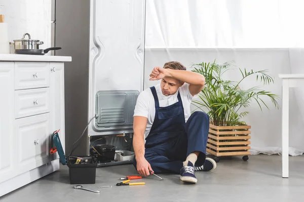 Tired adult repairman sitting on floor with hand on forehead and looking at tools while repairing refrigerator — Stock Photo