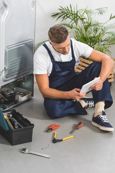 Erwachsener Reparateur sitzt auf dem Boden und benutzt digitales Tablet, während er Kühlschrank repariert — Stockfoto
