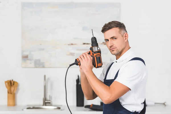 Lächelnder erwachsener Reparateur, der den Bohrer wie eine Waffe in der Hand hält und in die Kamera blickt — Stockfoto