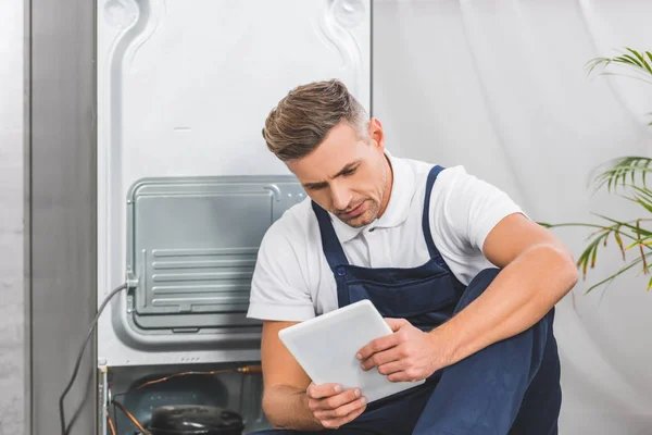 Erwachsener Reparateur sitzt auf dem Boden und benutzt digitales Tablet, während er Kühlschrank repariert — Stockfoto