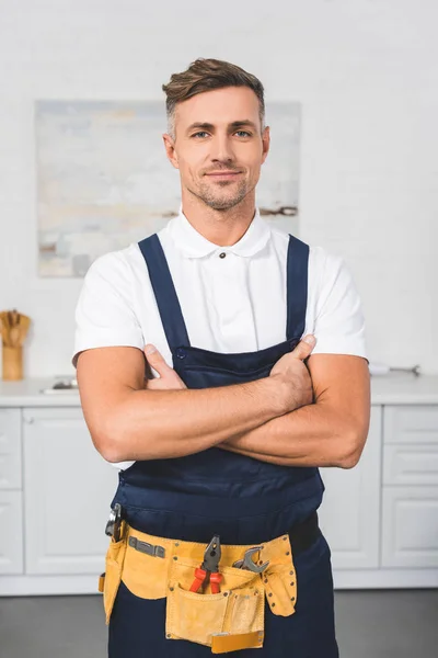 Reparador adulto no cinto de ferramentas de pé com os braços cruzados na cozinha e olhando para a câmera — Fotografia de Stock