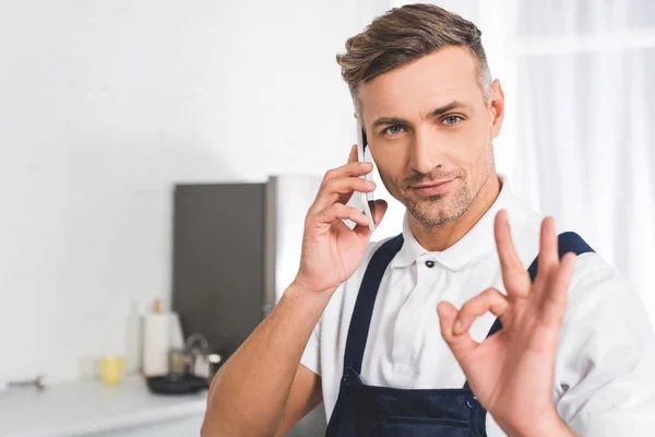 Reparador adulto hablando en el teléfono inteligente y mostrando signo ok - foto de stock