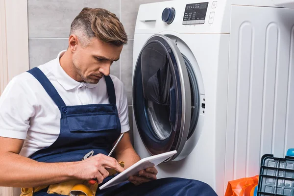 Réparateur adulte tenant tournevis et en utilisant une tablette numérique tout en réparant la machine à laver dans la salle de bain — Photo de stock