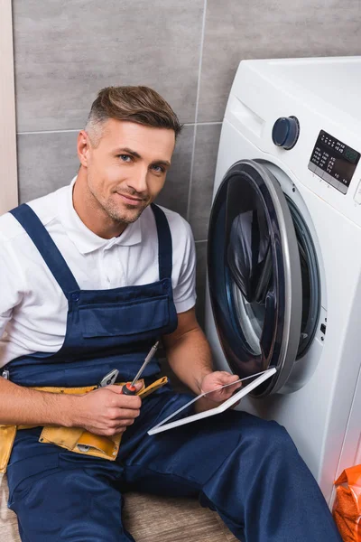 Erwachsener Reparateur mit Schraubenzieher und digitalem Tablet, während er Waschmaschine im Bad repariert und in die Kamera schaut — Stockfoto