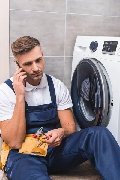 Adult repairman holding screwdriver and talking on smartphone while repairing washing machine in bathroom — Stock Photo