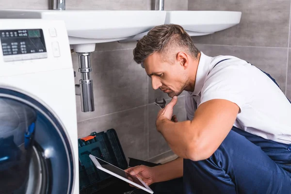 Nachdenklicher erwachsener Reparateur, der auf dem Boden sitzt und digitales Tablet benutzt, während er Waschbecken im Badezimmer repariert — Stockfoto