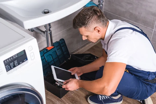 Erwachsener Reparateur sitzt auf dem Boden und benutzt digitales Tablet, während er Waschbecken im Badezimmer repariert — Stockfoto